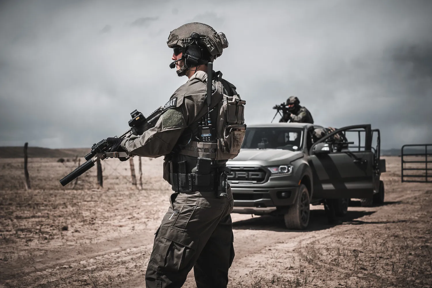 Operator wearing a helmet and headset, carrying a rifle and radio beside a pickup truck in the desert